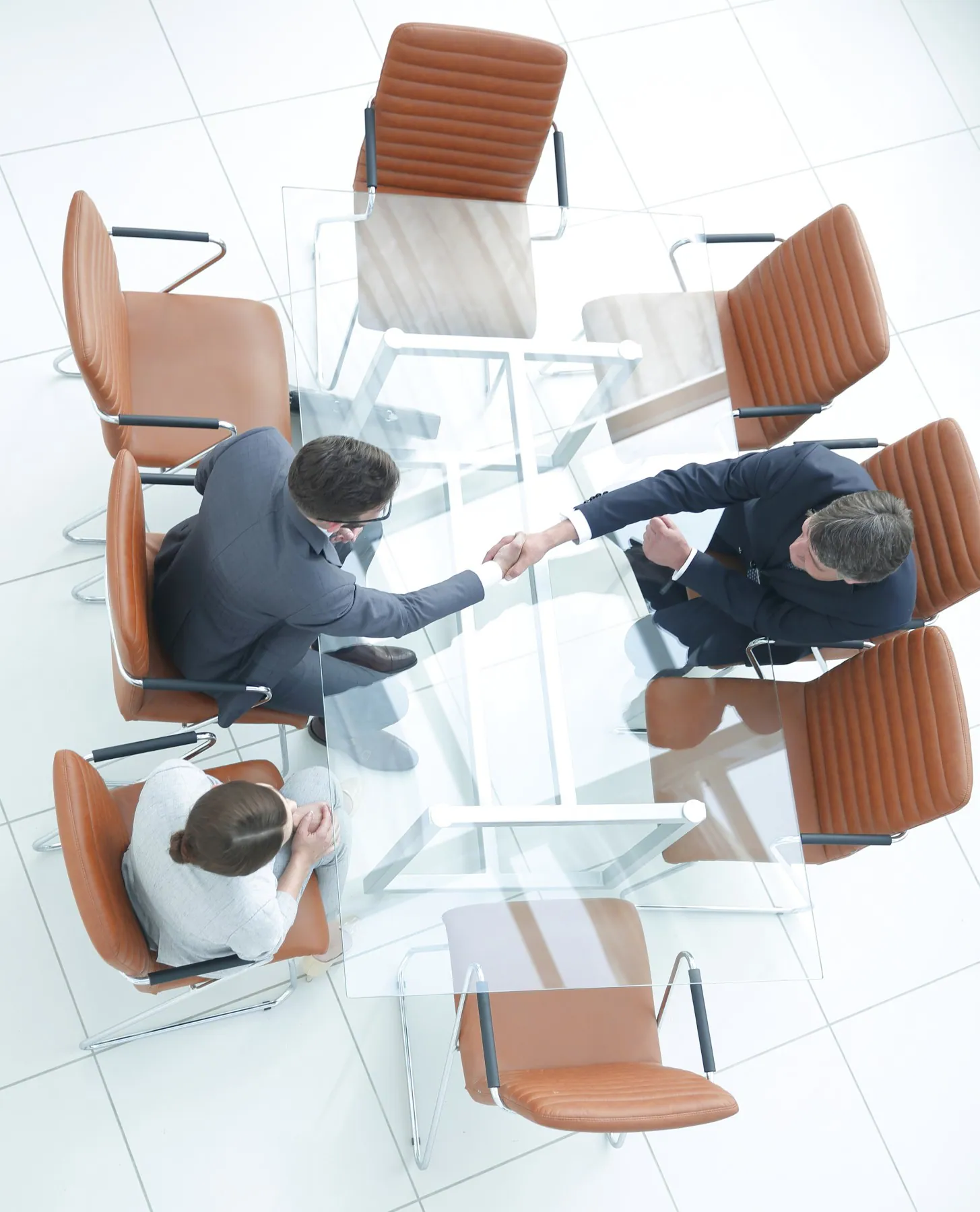 Three people in business attire sit around a glass conference table; two men shake hands while a woman looks on.
