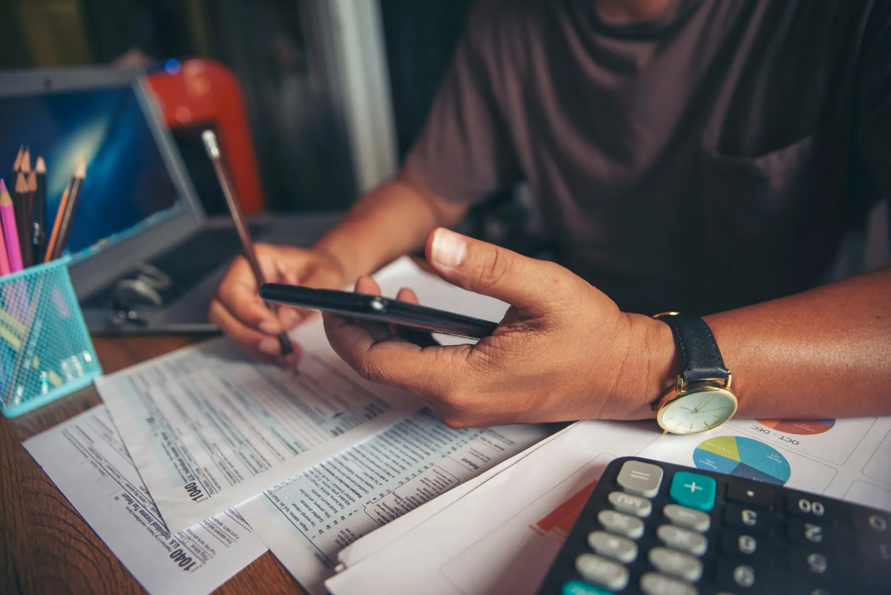 A person reviews documents and enters data on a smartphone, with a calculator, pencil holder, laptop, and charts visible on the desk.