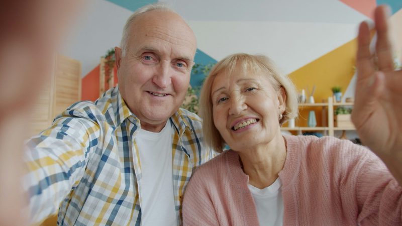 An older man and woman smile and wave at the camera while taking a selfie indoors.